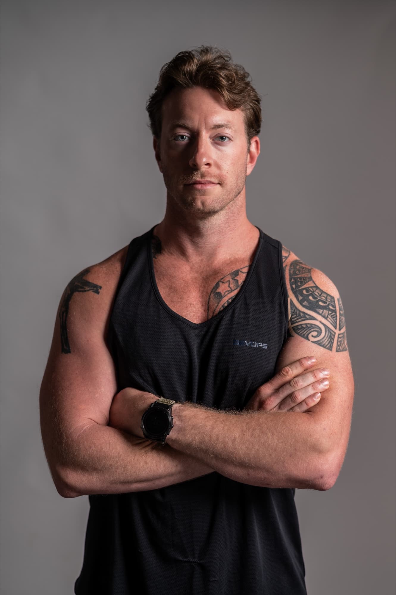 Austen Alexander in a black tank top with arms crossed, tattoos visible on both arms