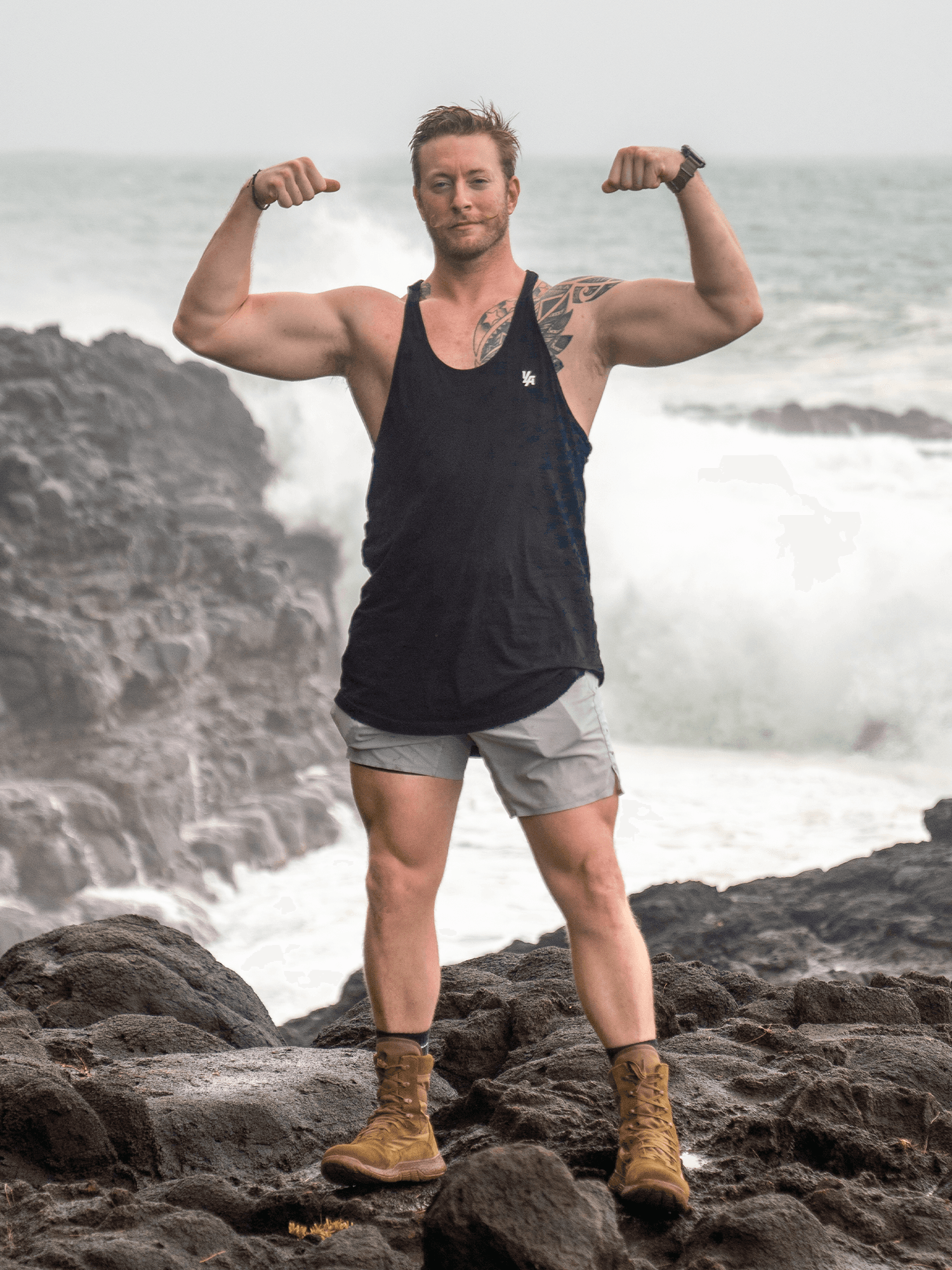 Austen Alexander flexing on a rocky coastline with waves crashing behind him