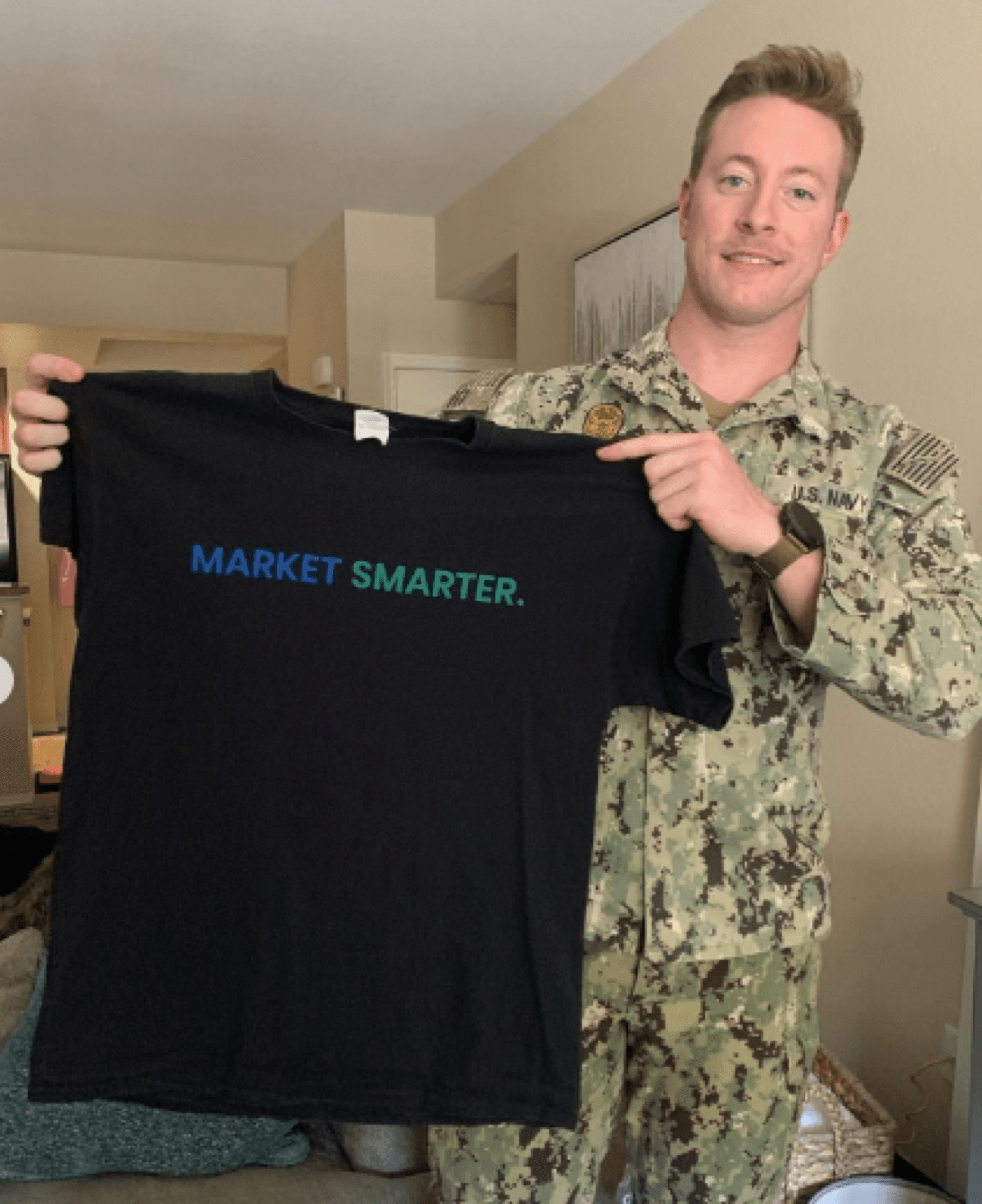 Austen Alexander in a Navy camouflage uniform holding up a black t-shirt printed Market Smarter